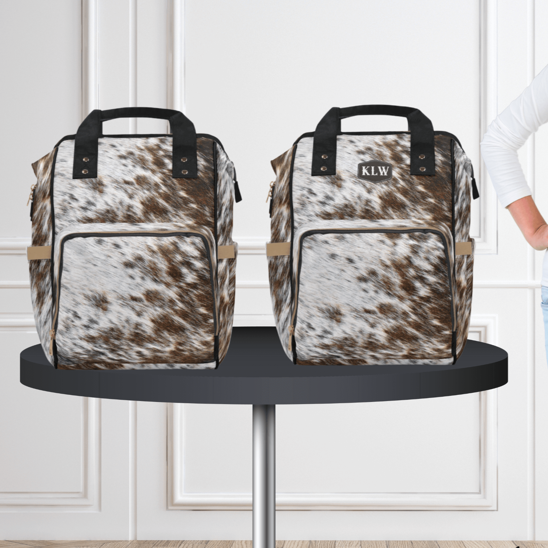 A teen girl stands next to a table with both the monogrammed cowhide diaper backpack and the plain cowhide diaper backpack.
