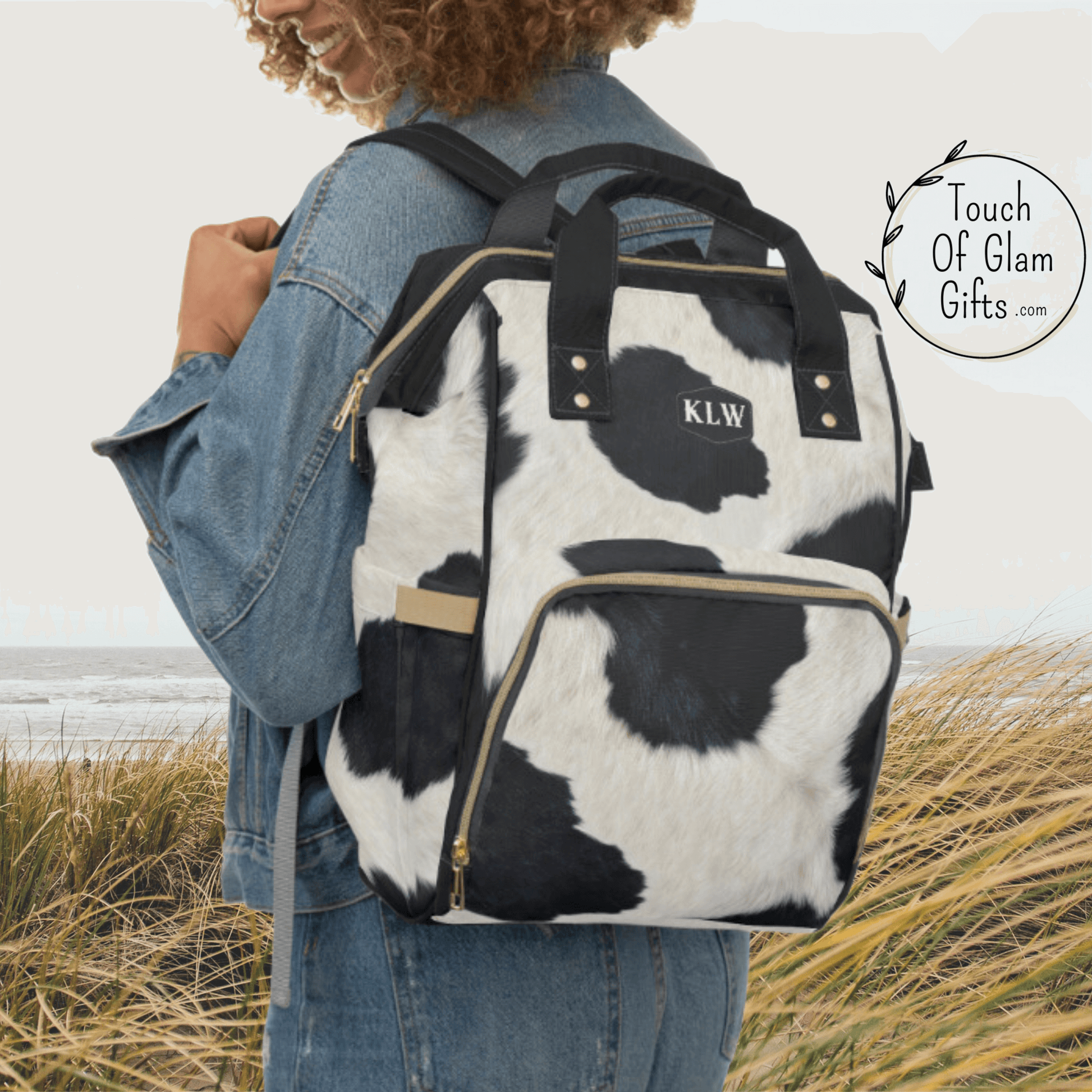 A cow print backpack is the perfect western travel gear for a cowgirl. THis backpack is shown on a female model wearing it over her shoulders with padded shoulder straps in black and has monogrammed initials.