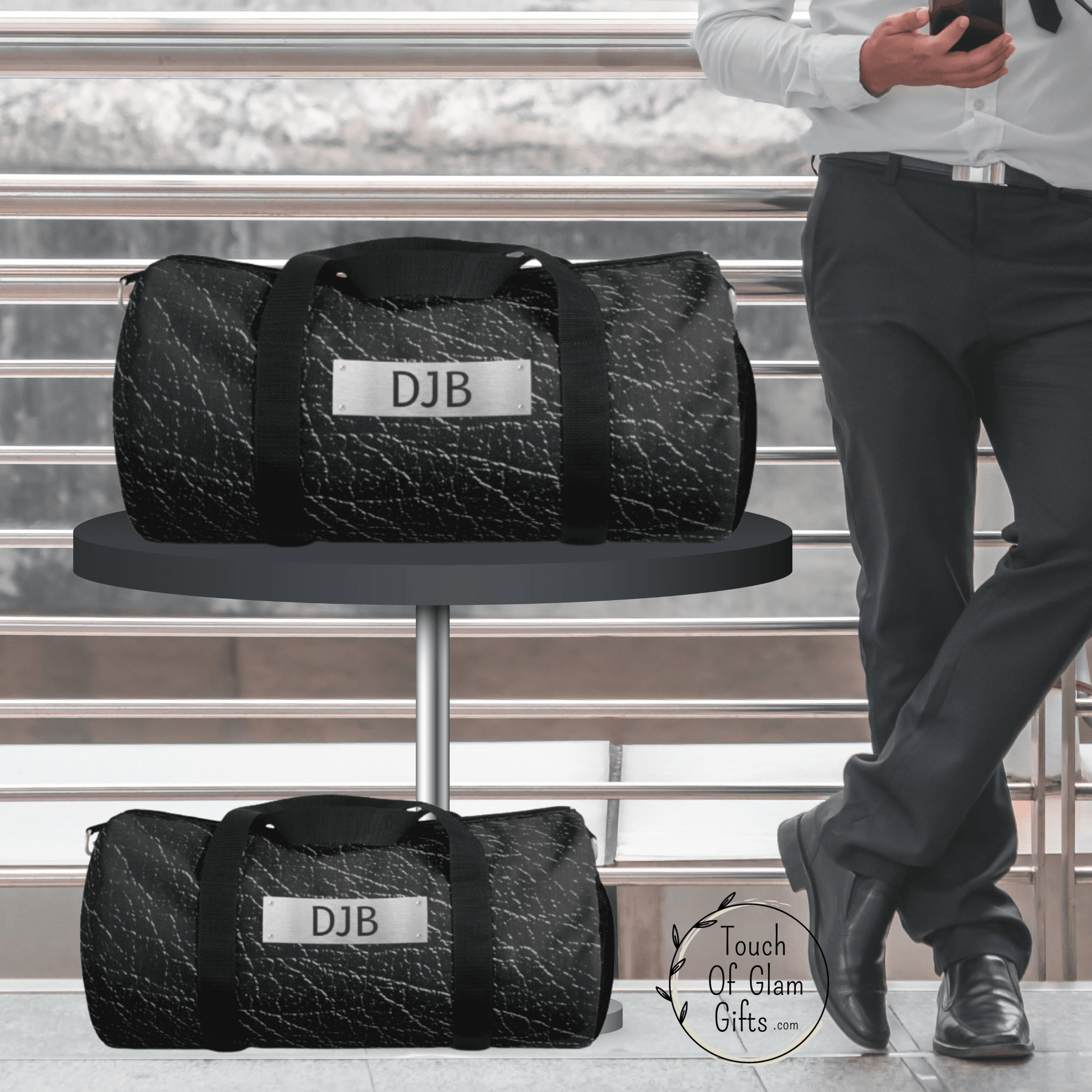 The large charcoal grey duffel bag sits on a table next to a guy checking his phone and the small duffel bag for guys is on the floor. both are identical and have monogrammed personalization for the perfect gift for guys.