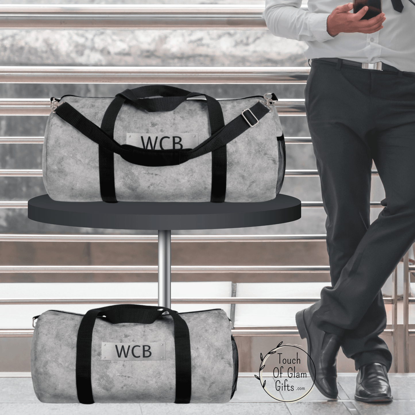 This duffel bag for men is the perfect gift for the business casual traveler. The large grey monogrammed duffel bag is on a table next to a man and the small carry on size duffel bag is on the floor.
